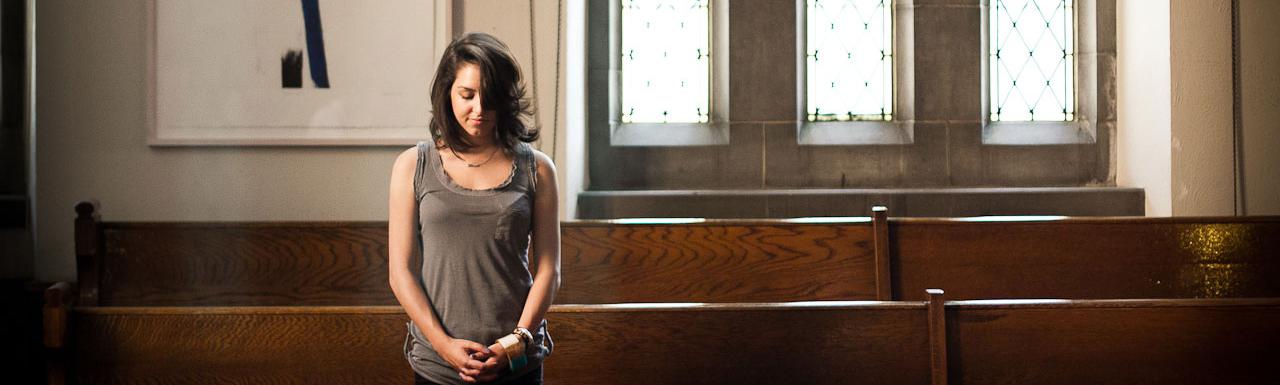 A young lady praying while standing in the pews of a church.